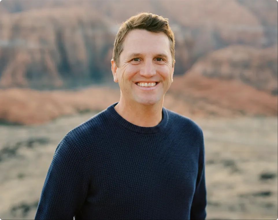 Matt smiling at the camera, wearing a dark blue sweater with a blurred canyon landscape in the background.