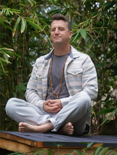 Matt meditating cross-legged and barefoot with his eyes closed, wearing a beaded necklace against a backdrop of dense green bamboo plants.