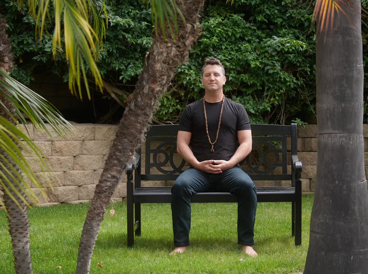Here is a short alt text for image_943c42.jpg: Matt sitting on a black bench with his eyes closed in meditation, wearing a black t-shirt and a long beaded necklace in a garden setting.