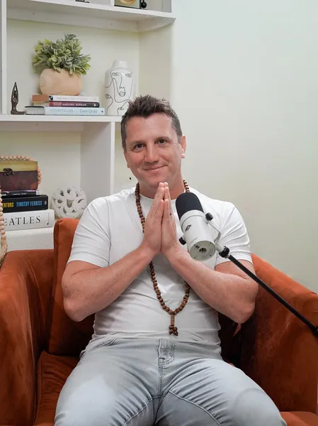 Matt sitting in an armchair with his hands in a prayer position, wearing a beaded necklace in front of a microphone.