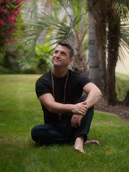 Matt sitting barefoot on the grass, looking upward with a slight smile in a tropical garden setting.