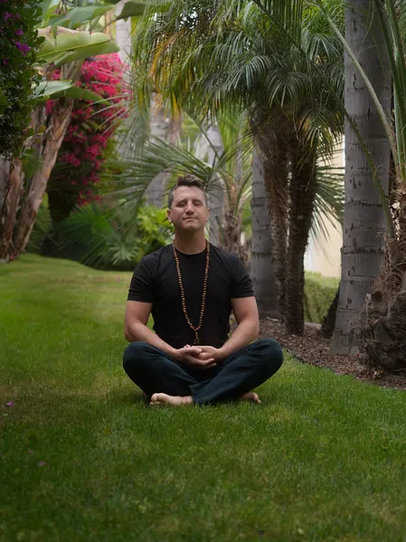 Matt meditating cross-legged on a lawn surrounded by tropical palm trees.