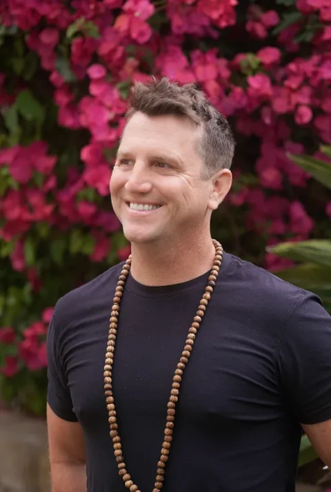 Matt smiling and looking to the side, wearing a black t-shirt and a long beaded necklace with vibrant pink bougainvillea flowers behind him.